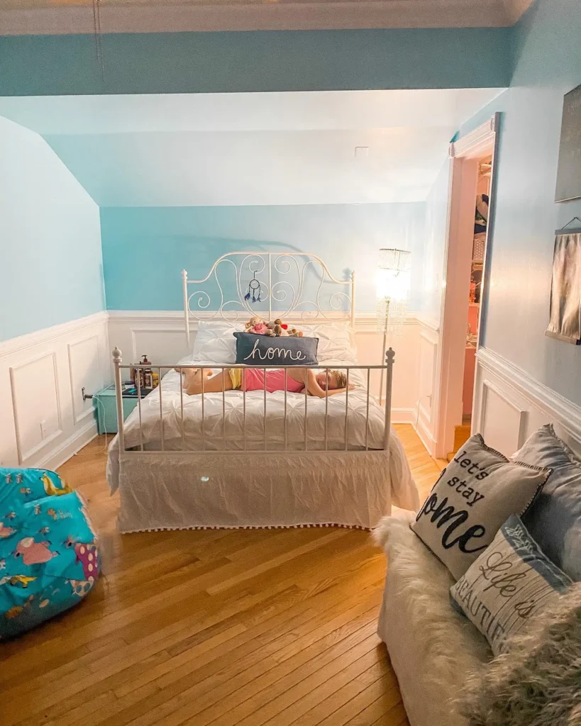 Preteen girls bedroom with aqua walls, white iron bed frame, wooden floors, and cozy accent pillows.
