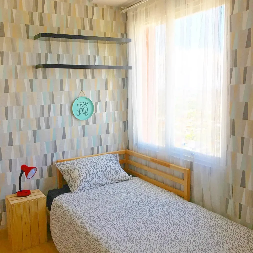 Compact guest bedroom with geometric wallpaper, single bed, and red lamp.