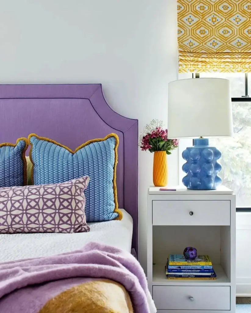 Preteen girls bedroom with purple headboard, patterned pillows, yellow curtains, and colorful lamp.
