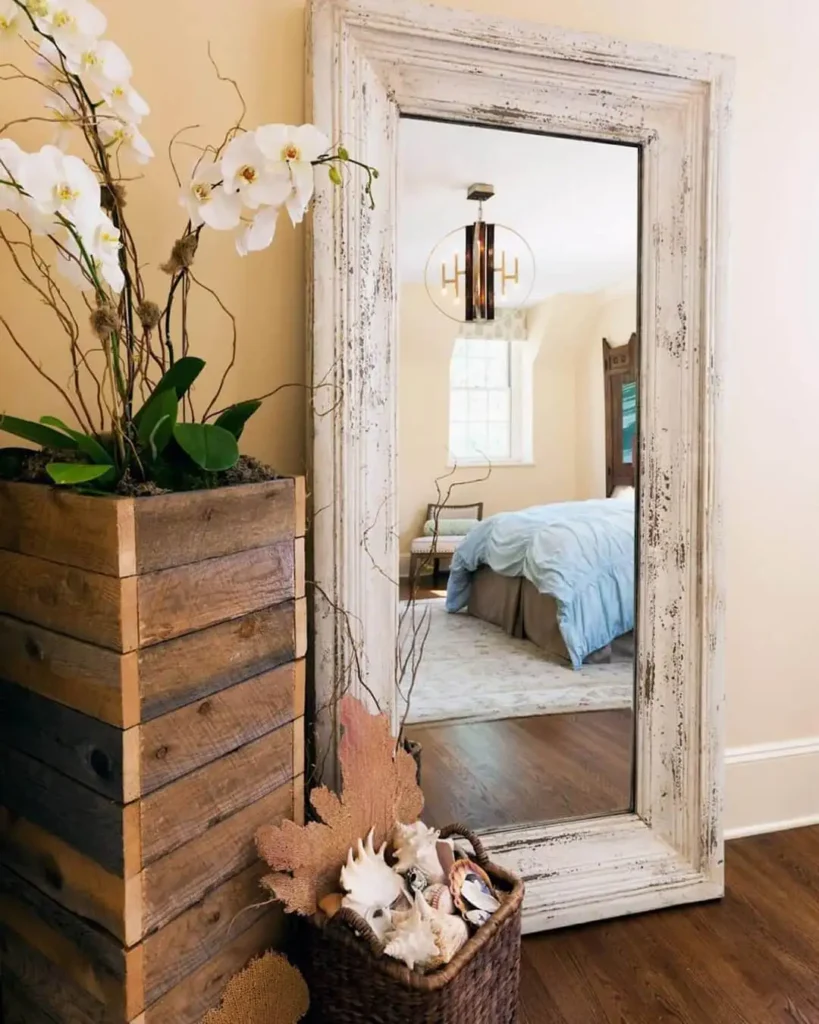 Distressed white frame full length mirror in bedroom with wood accents and floral arrangement.