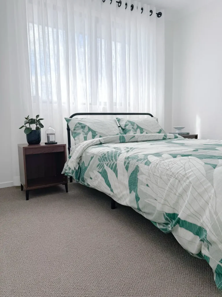 Guest bedroom with green leaf print bedding, sheer curtains, and dark wood nightstand.