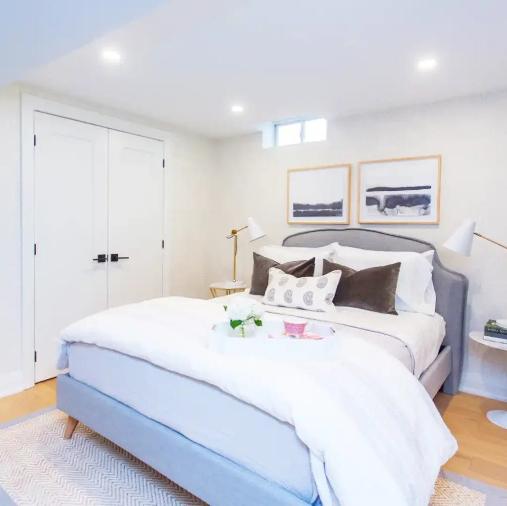 Basement guest bedroom with upholstered gray headboard, white bedding, and modern wall art.
