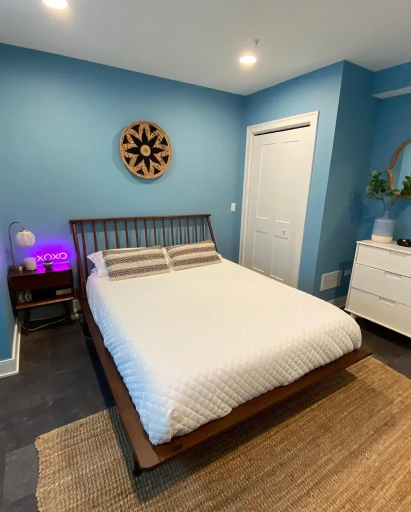 Basement bedroom with deep blue walls, neutral bed linens, and framed prints.
