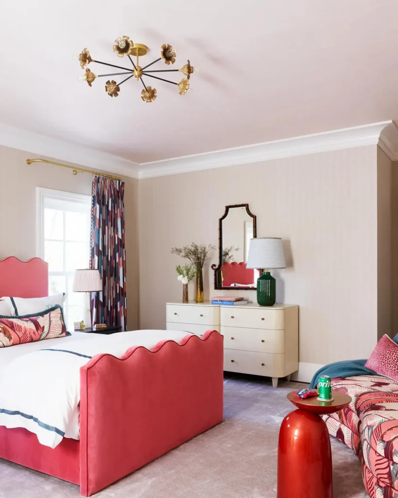 Coral upholstered bed with scalloped edges, patterned curtains, bold chandelier, and chic dresser.
