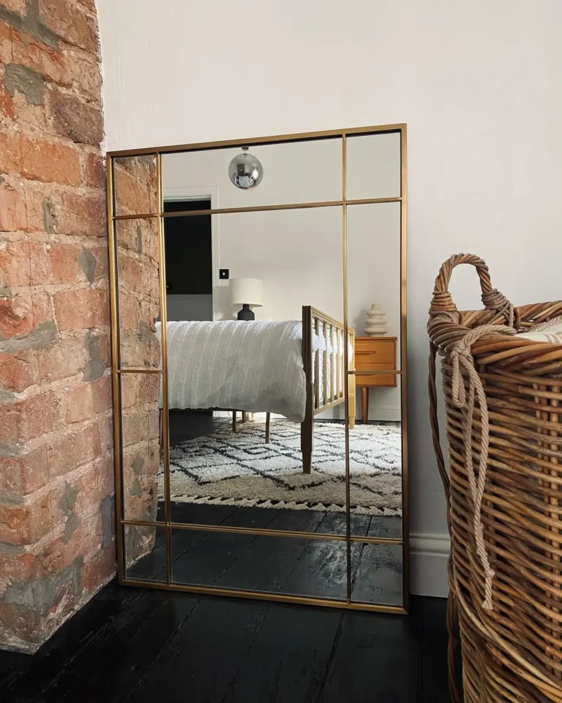 Full length mirror with gold grid design placed against brick wall in bedroom.