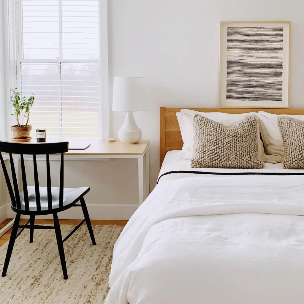 Guest bedroom office combo featuring white bedding, beige pillows, wooden desk, and abstract wall art in a minimalist layout.