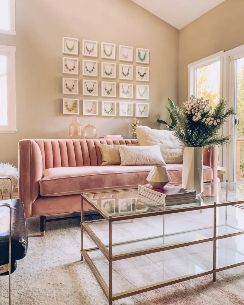 Blush pink sofa with gallery wall of framed necklaces and chic gold glass coffee table for feminine wall decor above couch.
