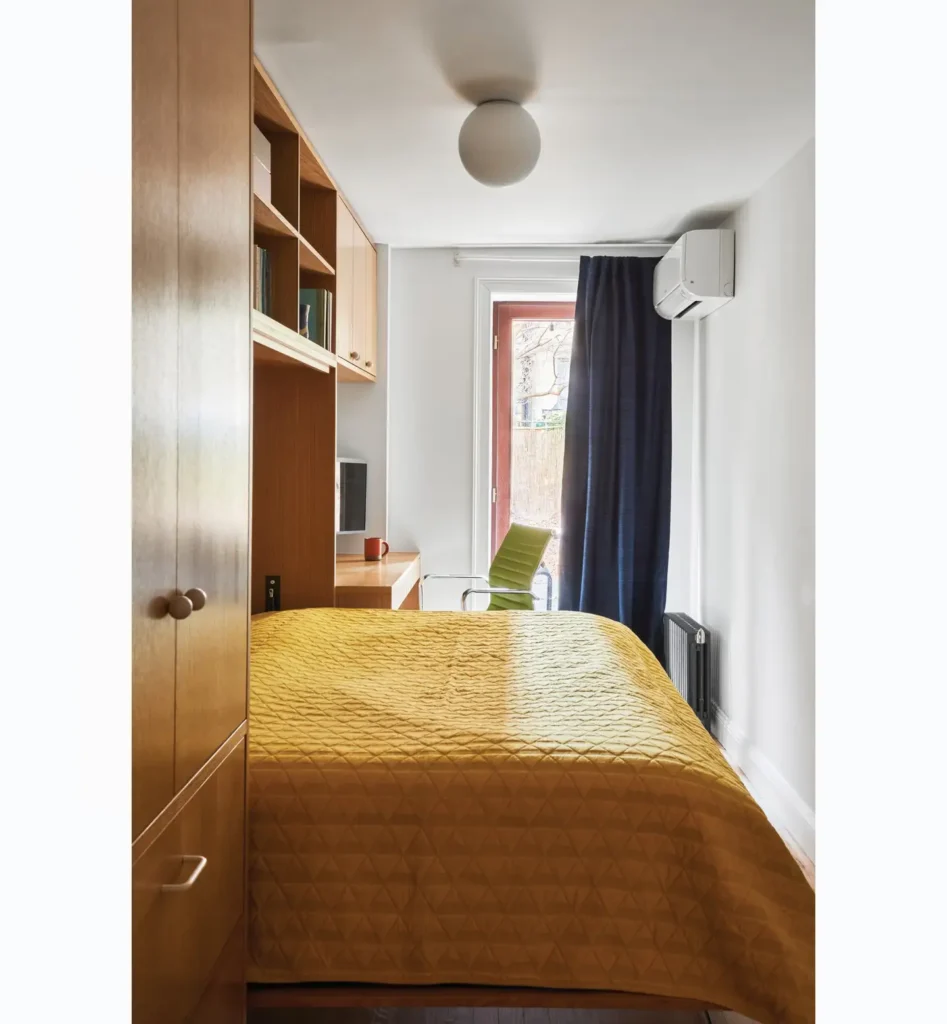 mustard bedding, green chair, built-in wardrobe, and desk near a window with blue curtains.