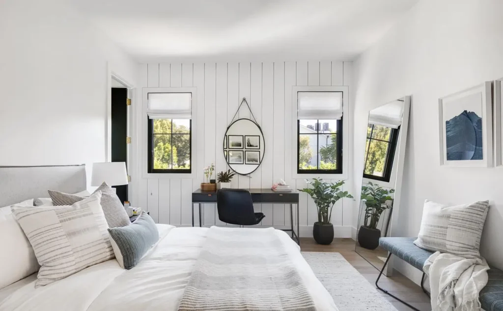 Guest bedroom office combo featuring black desk, round mirror, white and gray bedding, and dual windows with Roman shades.
